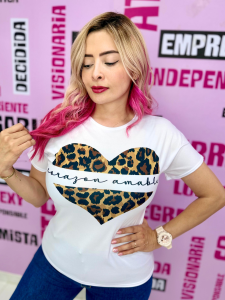 Mujer con cabello rubio y puntas rosadas, usando camiseta blanca con corazón de leopardo y la frase “corazón amable” al centro.