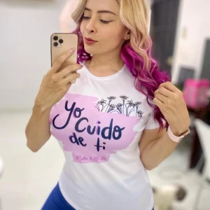 Mujer tomándose una selfie mientras usa una camiseta blanca con la frase “Yo cuido de ti – Mateo 6:28-30” y un diseño de flores negras sobre fondo rosado.