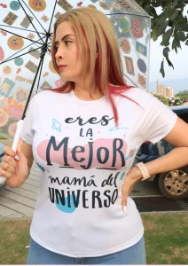 Mujer usando camiseta blanca con la frase “Eres la mejor mamá del universo”, parada al aire libre con una sombrilla decorada con ilustraciones coloridas.