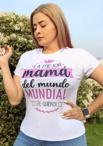 Mujer con camiseta blanca que dice “La mejor mamá del mundo mundial – ¡Te quiero!”, posando al aire libre con fondo de arbustos y cielo despejado.