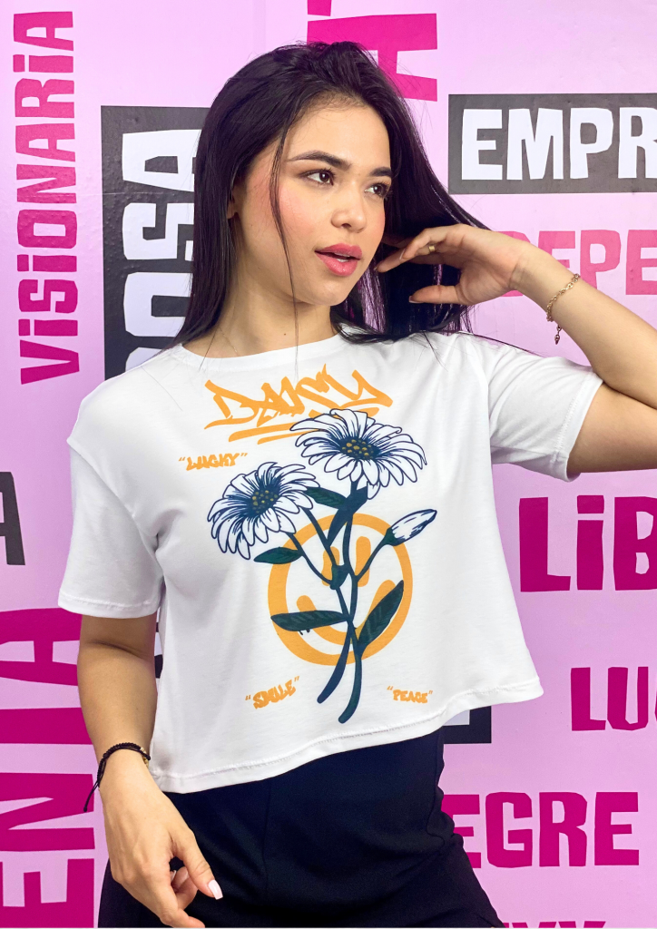Mujer con camiseta blanca ilustrada con margaritas y símbolos de paz, posando frente a un fondo rosa con palabras motivadoras.